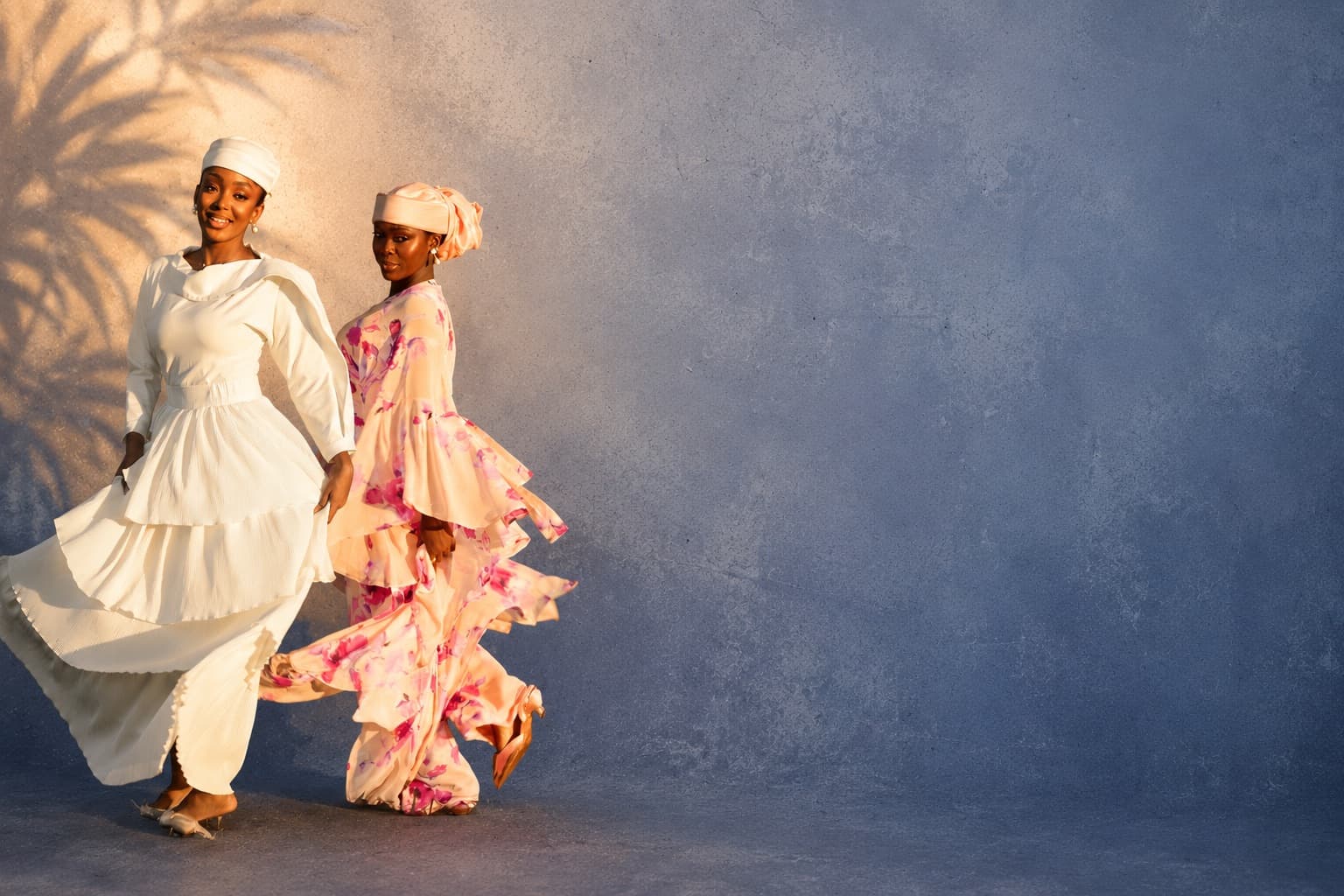 Two models in flowing ivory and blush floral dresses against a sunlit blue wall